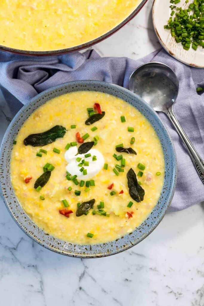 A speckled blue bowl filled with thick corn and bacon chowder is garnished with a dollop of sour cream, chopped chives, and crisp sage leaves on a marble table next to a silver ladle and a blue cloth napkin for corn and bacon chowder.