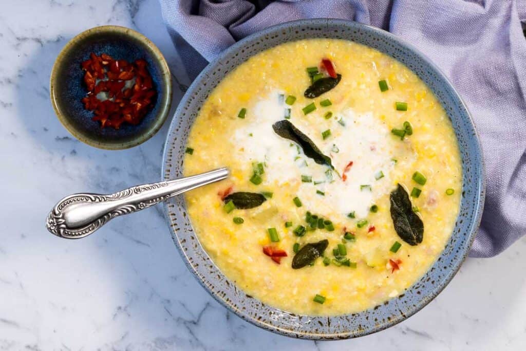 A close-up, overhead view of a blue ceramic bowl filled with corn and bacon chowder as the sour cream starts to swirl into the broth, topped with fried sage leaves and green onions next to a silver spoon and a small dish of diced red peppers for corn and bacon chowder.