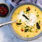 A close-up, overhead view of a blue ceramic bowl filled with corn and bacon chowder as the sour cream starts to swirl into the broth, topped with fried sage leaves and green onions next to a silver spoon and a small dish of diced red peppers for corn and bacon chowder.