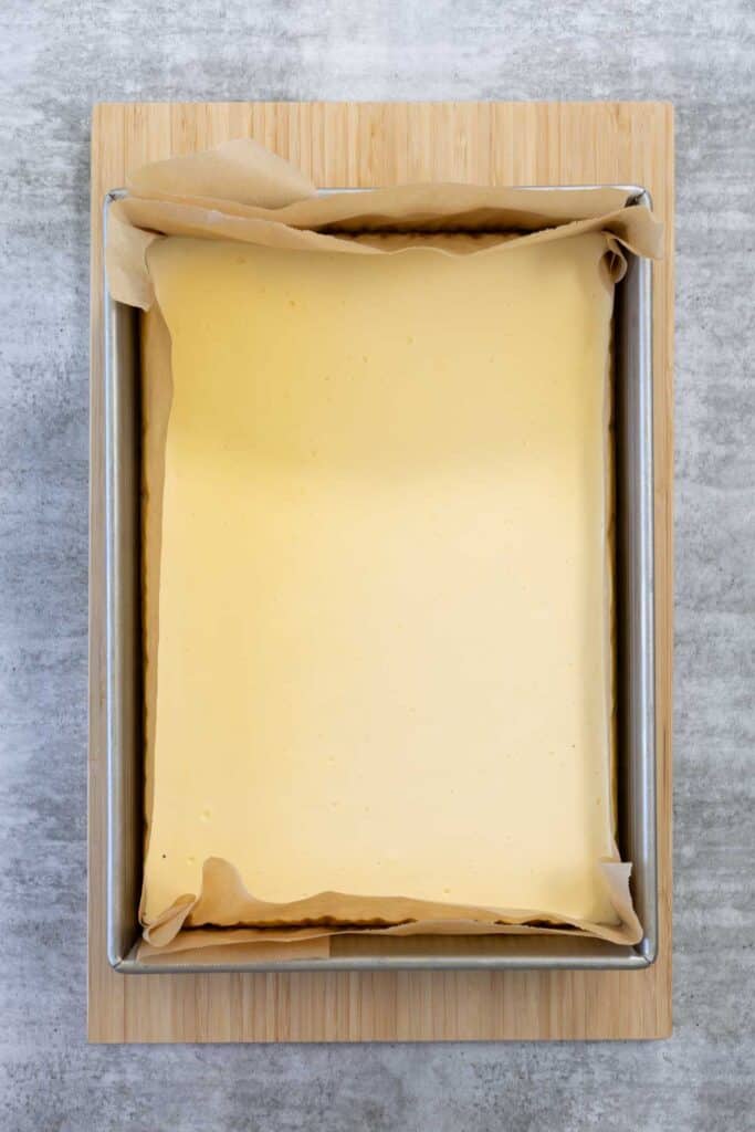 A baked, pale yellow Creme Brulee Cheesecake square, still in the parchment-lined metal pan, resting on a wooden cutting board to cool completely before chilling or slicing.