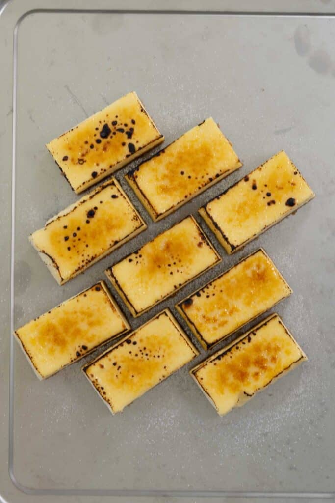 Nine rectangular slices of Creme Brulee Cheesecake arranged on a baking sheet, each topped with a thin layer of granulated sugar that has been caramelized and browned using a kitchen torch.