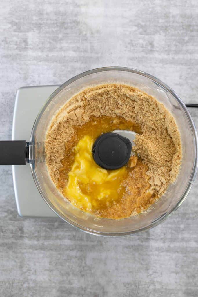 Golden-brown biscuit crumbs and a pool of melted butter combined in the bowl of a food processor, ready to be pulsed to form the base crust for the Creme Brulee Cheesecake.