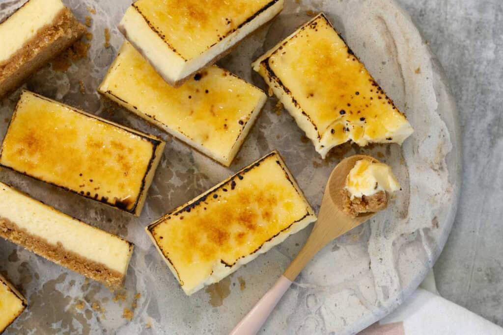An overhead close-up of several finished Creme Brulee Cheesecake bars—some plain and some torched—arranged on a marble serving board, with a small wooden spoon holding a bite-sized sample.