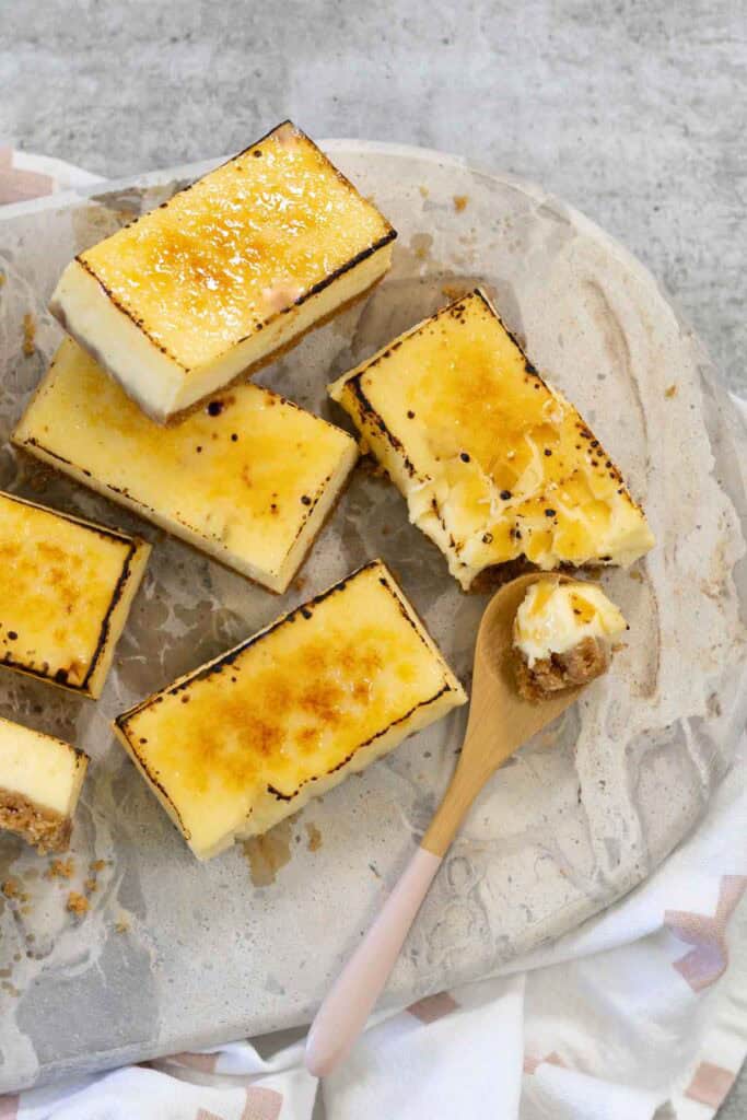 A top-down, vertical view of six finished rectangular Creme Brulee Cheesecake bars, four with caramelized sugar tops, displayed on a light gray marble serving platter with a wooden spoon holding a piece of crust and filling.