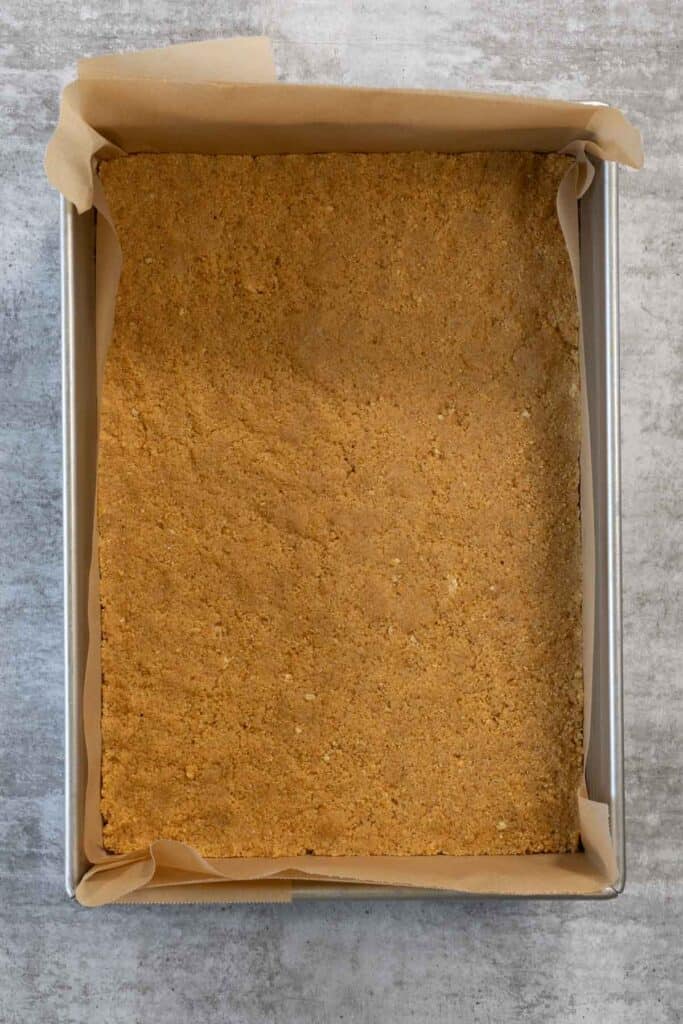 A finished, compacted biscuit crust is pressed firmly into a flat, even layer spanning the bottom of a square baking pan lined with parchment paper for the Creme Brulee Cheesecake.