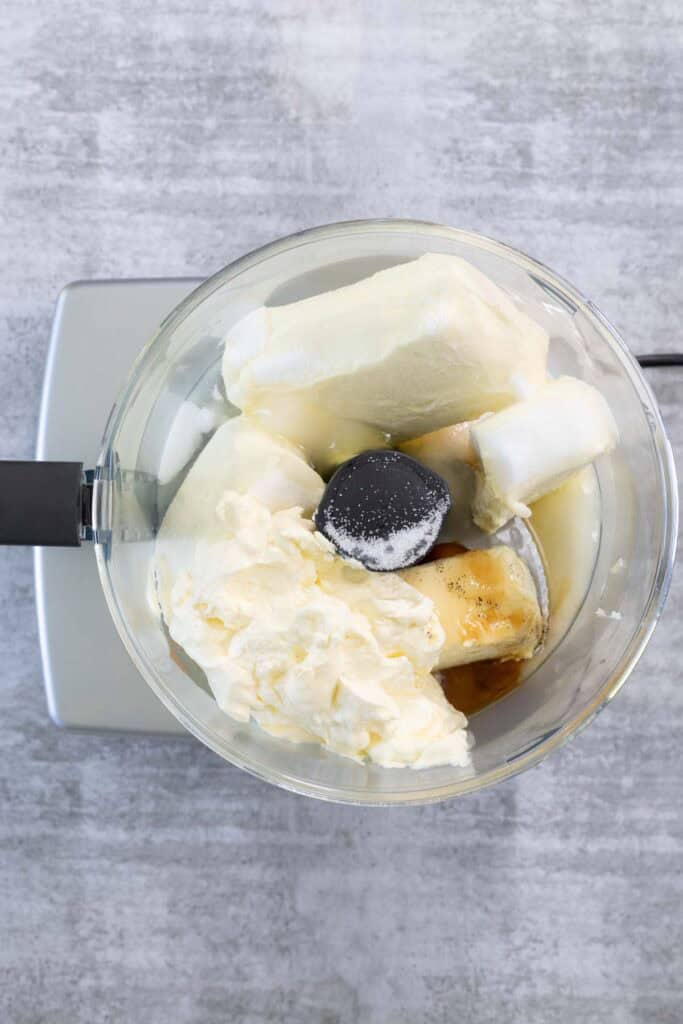 Large blocks of cream cheese, scoops of heavy cream, sugar, and vanilla extract are placed together inside the bowl of a food processor, ready to be blended into the filling for the Creme Brulee Cheesecake.