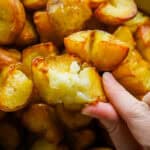 Two fingers hold a crispy roasted potato up to the camera from below. The potato has been broken open to show the crispy exterior and the fluffy, soft interior. Underneath the potato a bowl of roasted potatoes can be seen slightly out of focus.