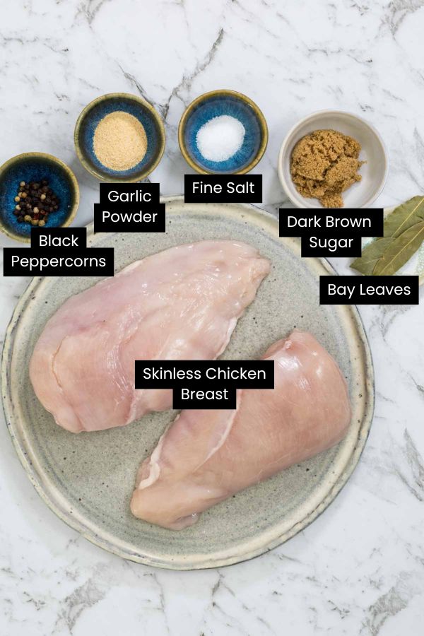 Two raw, boneless chicken breasts are laid out on a large ceramic plate surrounded by small bowls of brown sugar, kosher salt, garlic powder, whole peppercorns, and bay leaves on a white marble surface for dry brined chicken.
