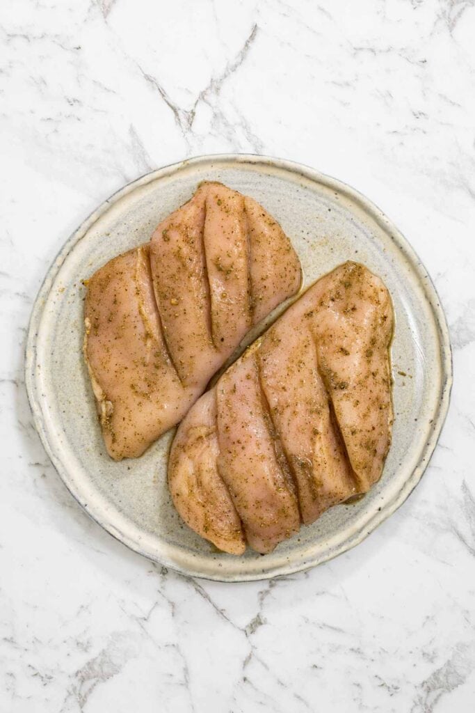 Two raw chicken breasts are scored with shallow diagonal cuts and thoroughly coated in a brown spice rub on a round ceramic plate for dry brined chicken.