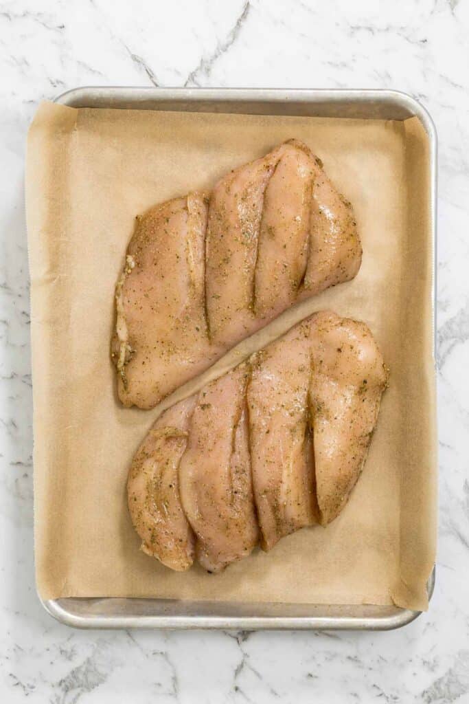 Two seasoned, scored raw chicken breasts are transferred onto a silver baking sheet lined with brown parchment paper for dry brined chicken.