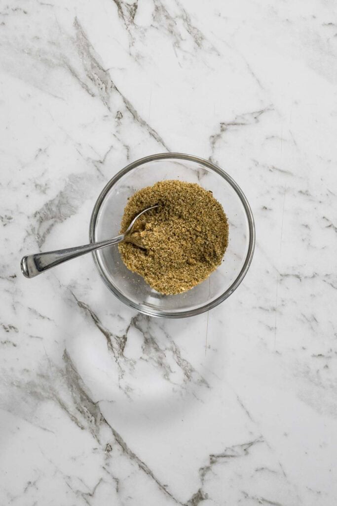 A small glass bowl filled with a mixed brown and green dry brine seasoning rub sits on a marble countertop with a silver spoon resting inside for dry brined chicken.
