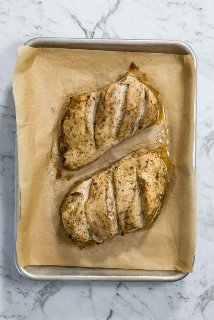 Two baked, golden-brown chicken breasts with clear score marks sit on a parchment-lined sheet pan surrounded by natural juices for dry brined chicken.