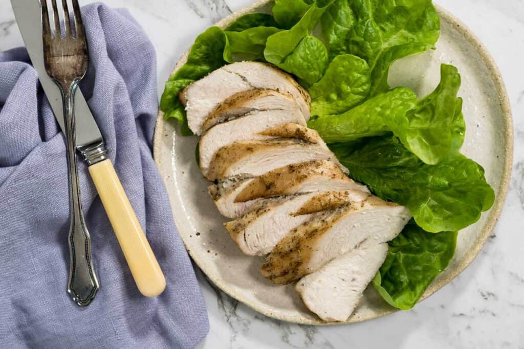 Slices of juicy roasted chicken breast are arranged on a light ceramic plate alongside fresh green butter lettuce leaves, complete with a silver fork and knife on a blue cloth napkin for dry brined chicken.