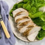 Slices of juicy roasted chicken breast are arranged on a light ceramic plate alongside fresh green butter lettuce leaves, complete with a silver fork and knife on a blue cloth napkin for dry brined chicken.