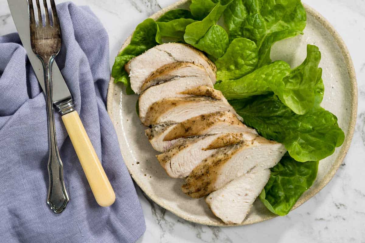 Slices of juicy roasted chicken breast are arranged on a light ceramic plate alongside fresh green butter lettuce leaves, complete with a silver fork and knife on a blue cloth napkin for dry brined chicken.