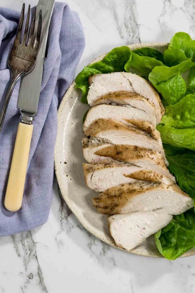 A close-up, top-down view of sliced roasted chicken breast overlapping on a ceramic plate next to green lettuce leaves, with a vintage-style knife and fork resting on a purple napkin to the left for dry brined chicken.