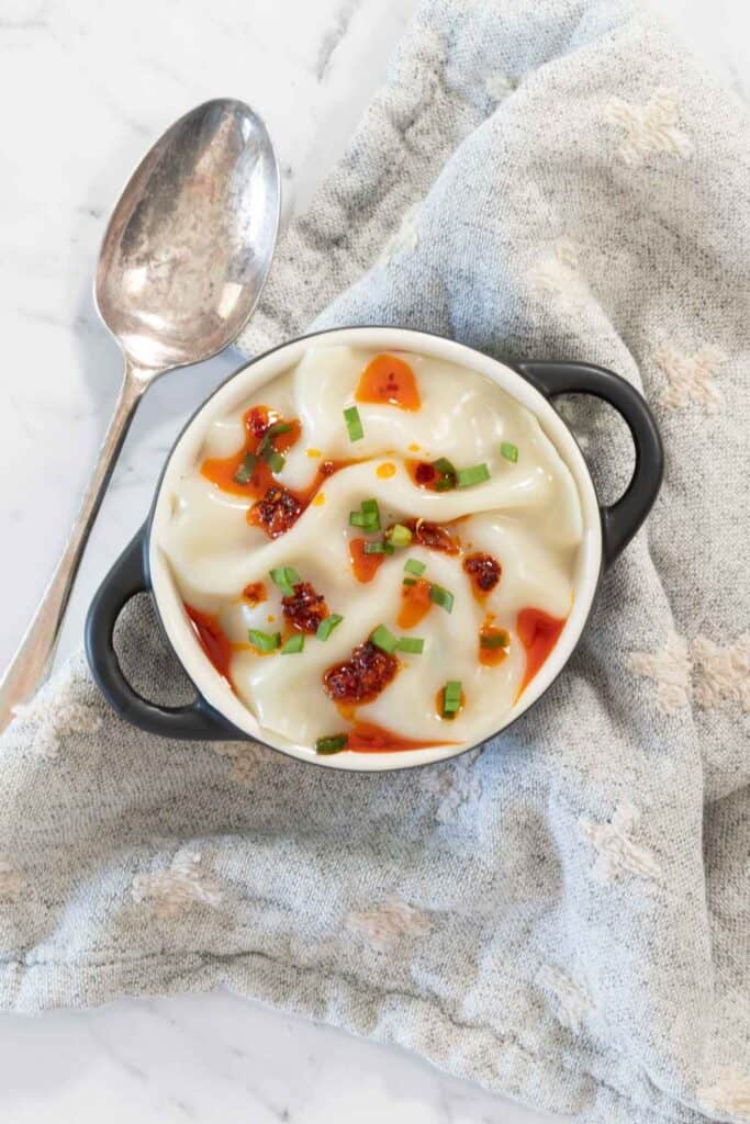 Overhead view of a cooked dumpling lasagne in a small black-handled cocotte with a silver spoon resting on a gray patterned cloth.