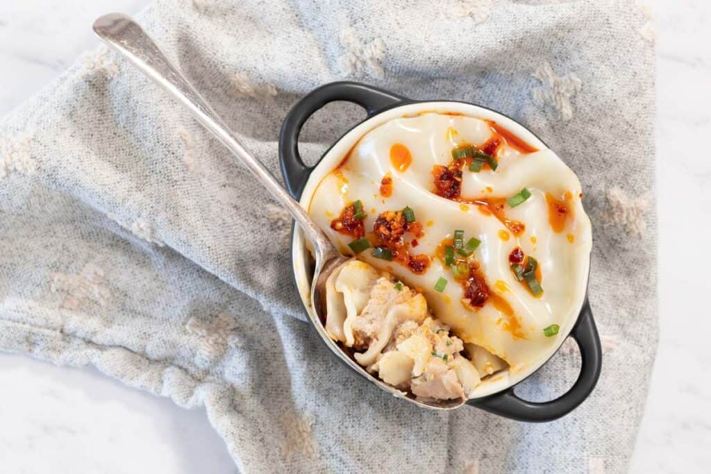 Overhead view of a cooked dumpling lasagne in a small black-handled cocotte topped with chili crisp and sliced green onions on a blue patterned cloth.