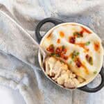 Overhead view of a cooked dumpling lasagne in a small black-handled cocotte topped with chili crisp and sliced green onions on a blue patterned cloth.