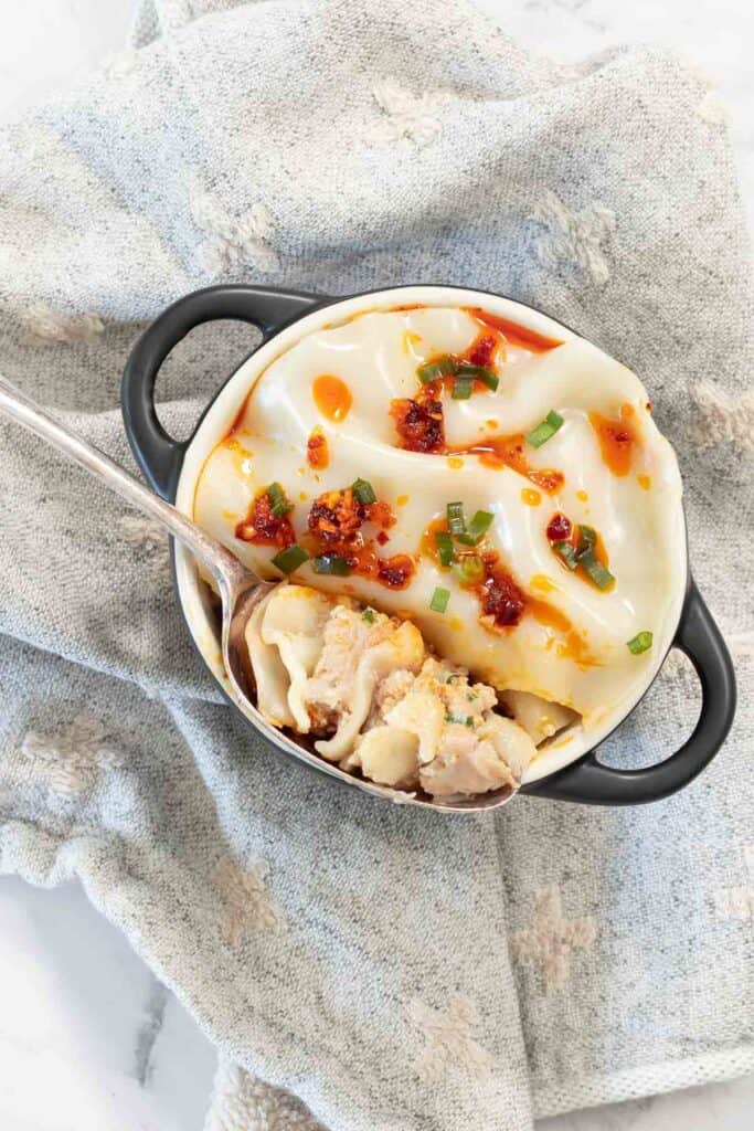 Silver spoon scoops into a cooked dumpling lasagne topped with chili crisp and sliced green onions inside a small black-handled cocotte.