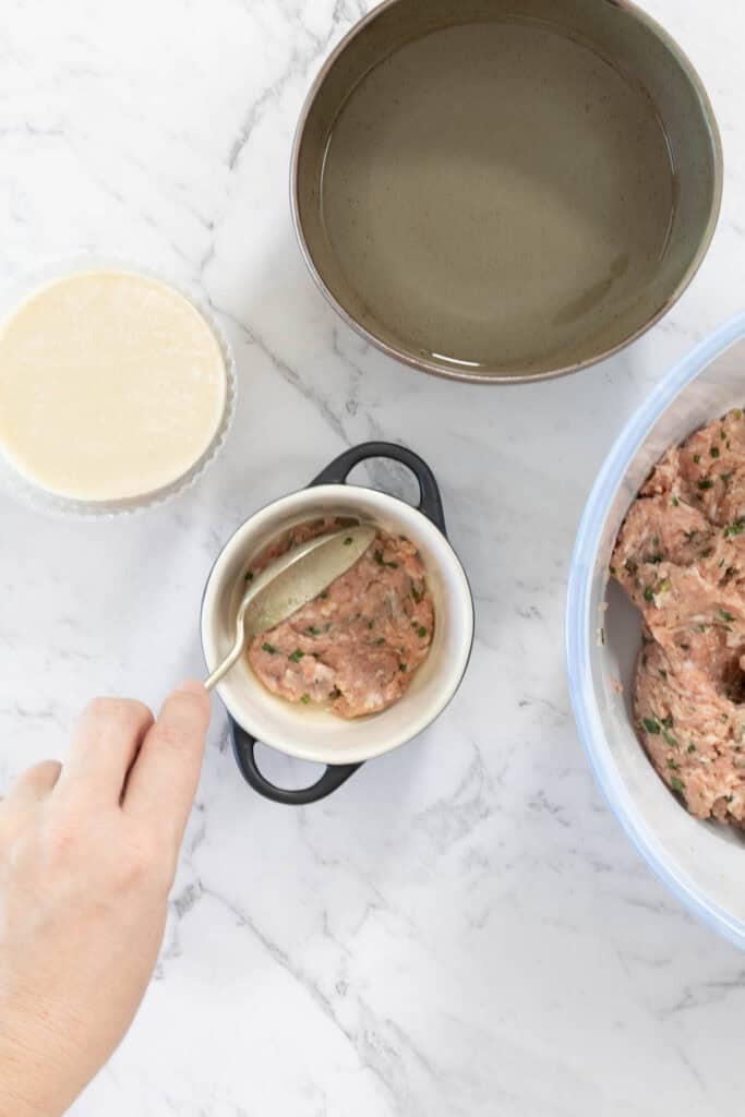 Metal spoon presses a flat layer of seasoned ground pork into a small white ceramic baking dish for dumpling lasagne.