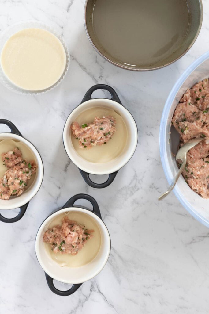 Small portions of seasoned ground pork are placed onto round dumpling wrappers inside three individual white cocottes for dumpling lasagne.