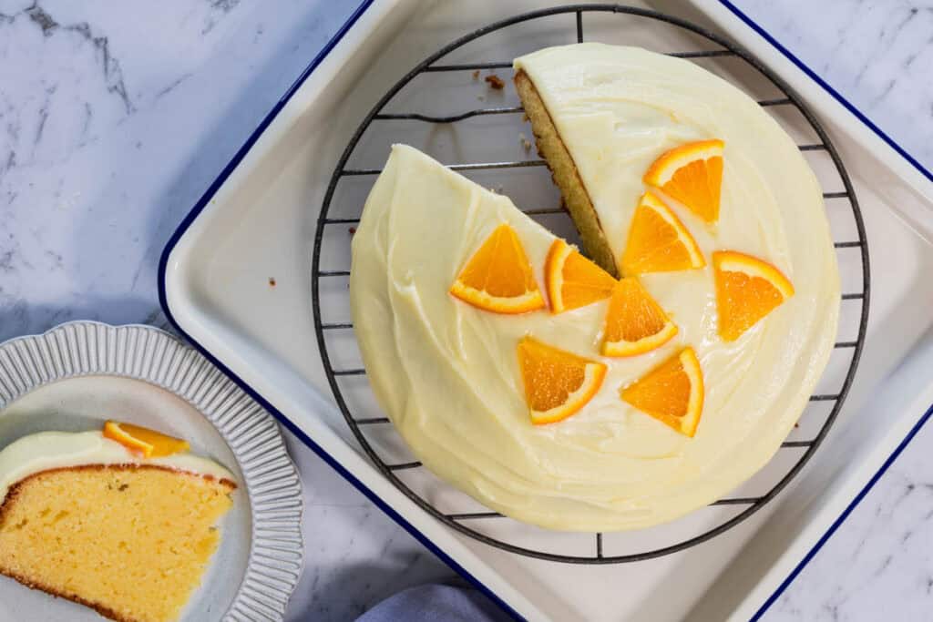 Sliced sponge cake topped with smooth white frosting and fresh orange wedges sits on a wire rack next to a single serving on a fluted plate for easy orange cake.