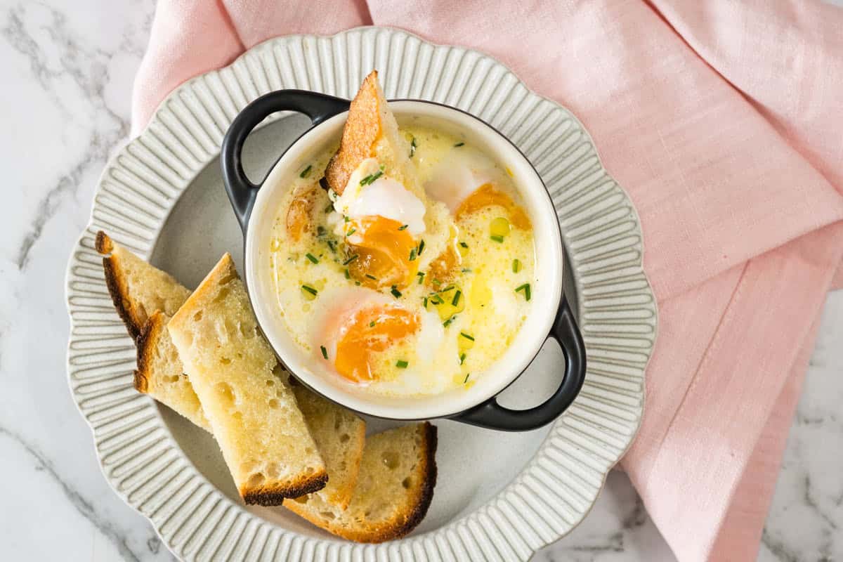 steam-baked eggs topped with chives in a small black ramekin, served with toasted sourdough soldiers on a ribbed plate alongside a pink napkin.