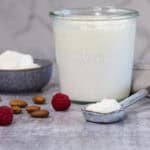 A small grey bowl with plain yoghurt in it; next to the bowl is a larger glass jar of yoghurt, and a ceramic spoon. There are almonds and raspberries scattered on the counter nearby.