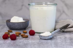 A small grey bowl with plain yoghurt in it; next to the bowl is a larger glass jar of yoghurt, and a ceramic spoon. There are almonds and raspberries scattered on the counter nearby.