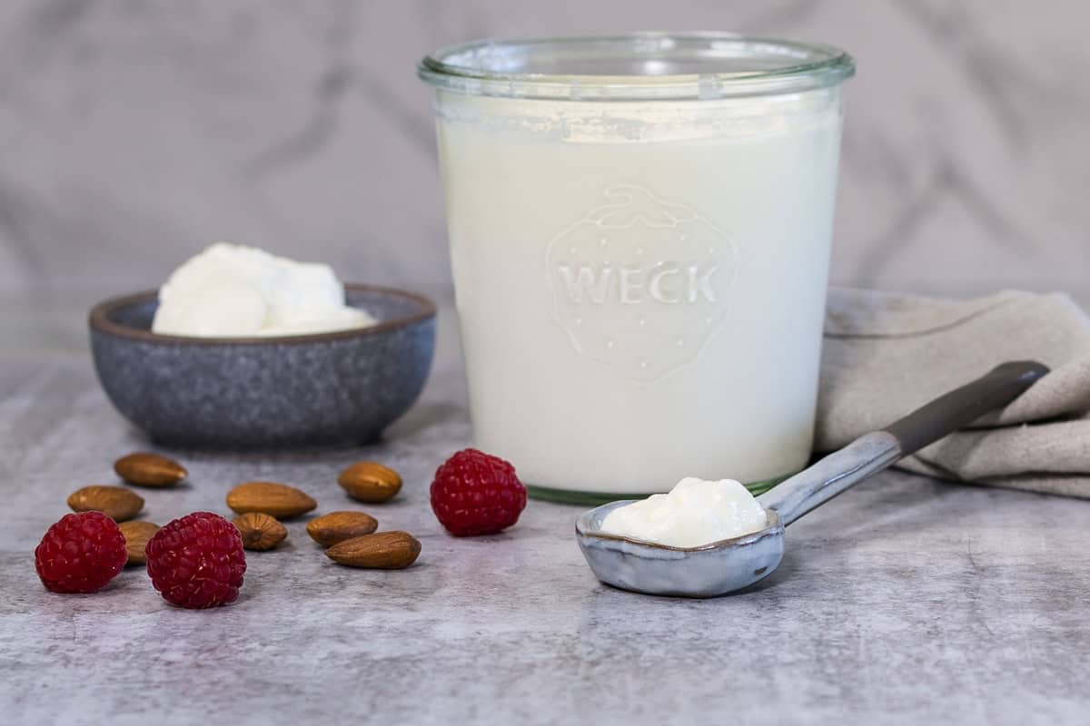 A small grey bowl with plain yoghurt in it; next to the bowl is a larger glass jar of yoghurt, and a ceramic spoon. There are almonds and raspberries scattered on the counter nearby.