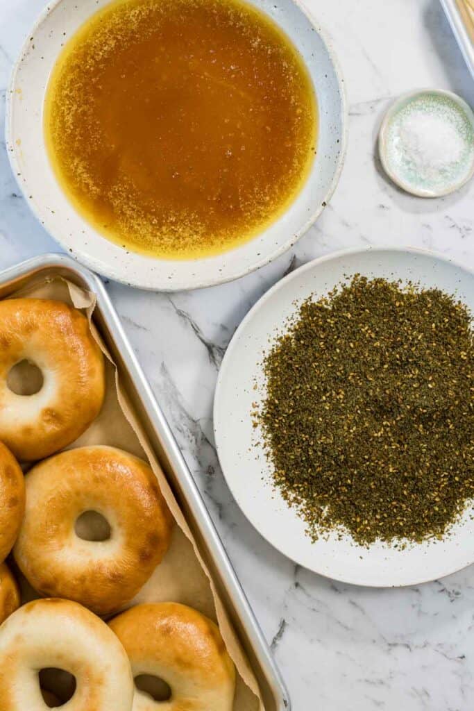 A tray of golden baked bagels sits next to a bowl of melted honey butter and a plate of green zaatar spice blend for zaatar bagels.