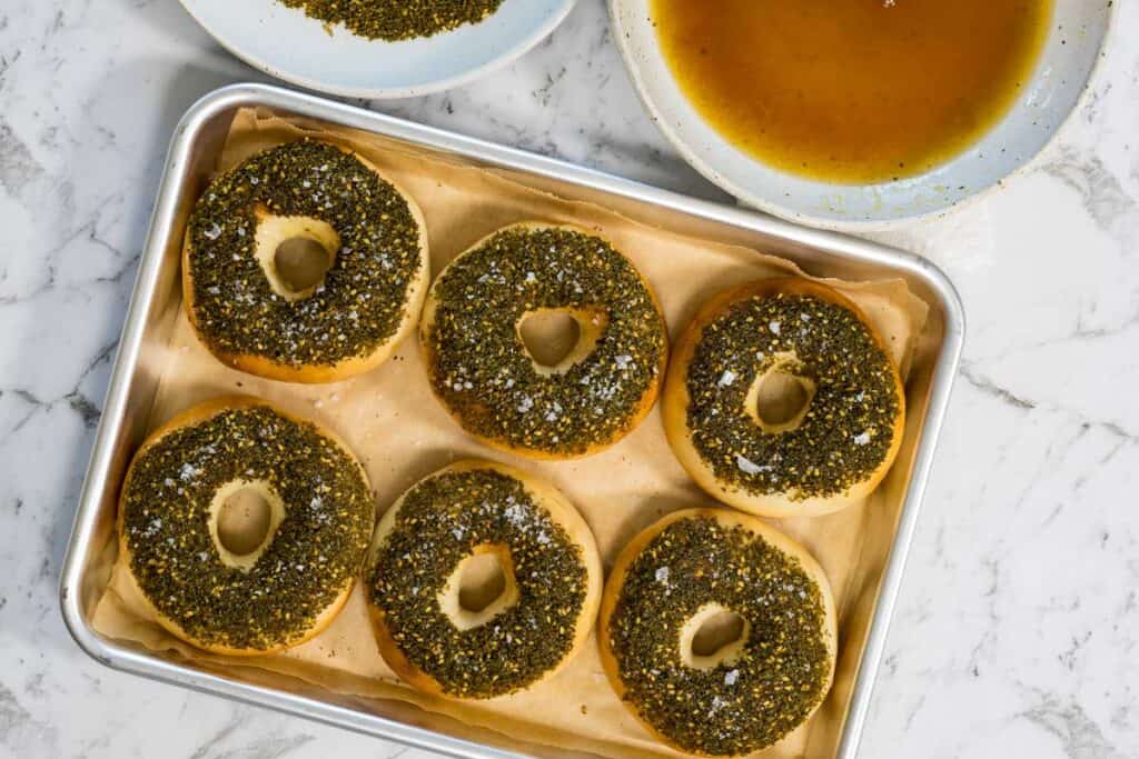 Six golden bagels topped with a thick layer of green zaatar herbs and sea salt flakes are arranged on a parchment-lined baking sheet for zaatar bagels.