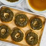 Six golden bagels topped with a thick layer of green zaatar herbs and sea salt flakes are arranged on a parchment-lined baking sheet for zaatar bagels.