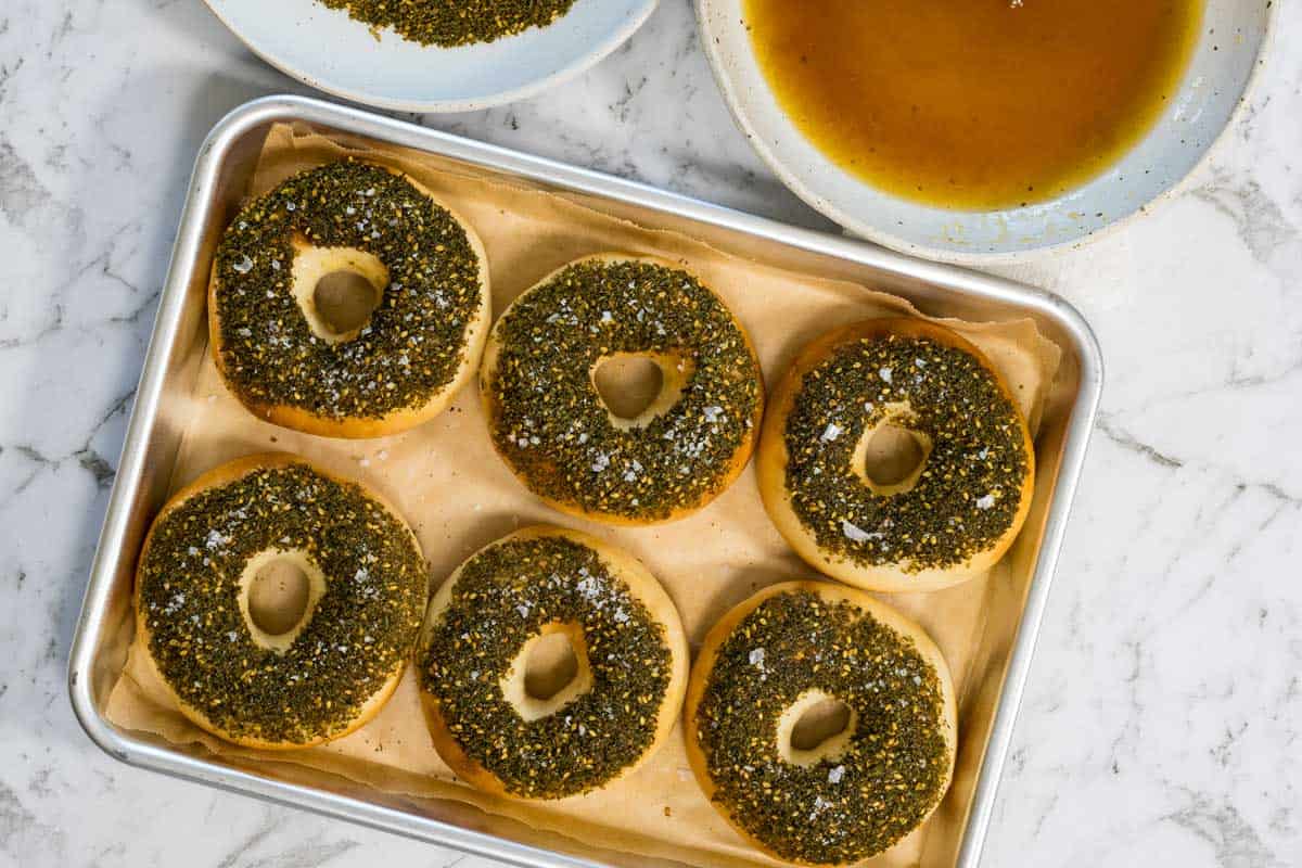Six golden bagels topped with a thick layer of green zaatar herbs and sea salt flakes are arranged on a parchment-lined baking sheet for zaatar bagels.