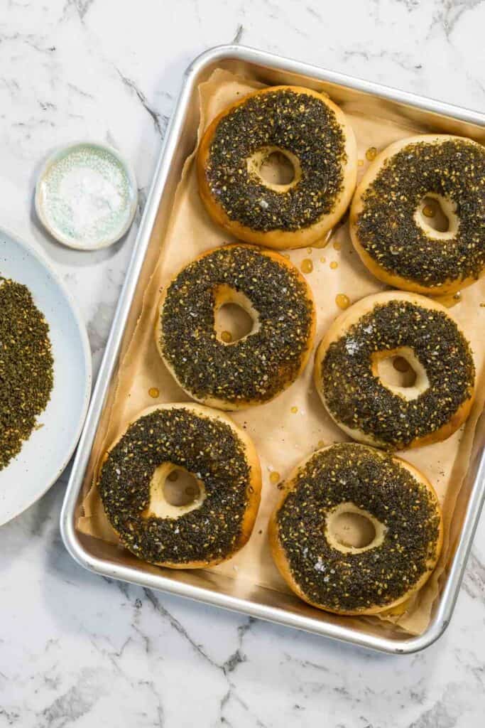 A final tray of finished bagels is drizzled with extra golden honey, highlighting the zaatar bagels alongside bowls of extra spices and sea salt.