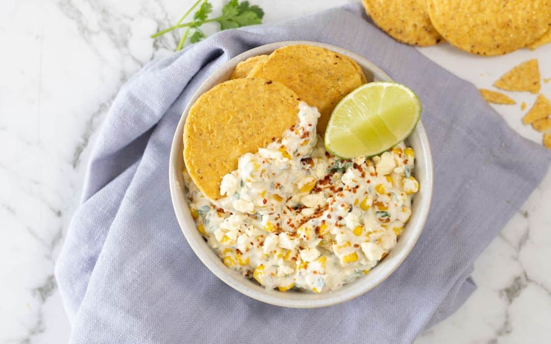 creamy corn appetizer topped with crumbled cheese, chili seasoning, and a fresh lime wedge, served with round tortilla chips in a ceramic bowl over a blue linen napkin
