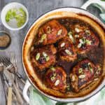 A round white cast iron baking dish is viewed from above. In the dish are 6 roasted chicken cutlets with miso maple marinade. There are sesame seeds and scallions sprinkled on top, and next to the dish is a pile of cutlery and extra scallions and seeds.