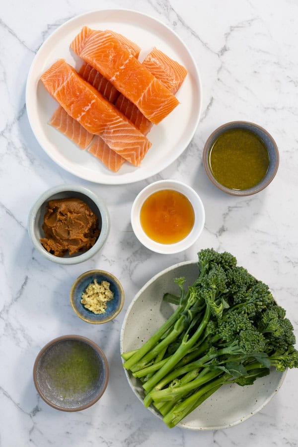 Ingredients for roasted salmon including raw fillets, broccolini, miso paste, ginger, honey, and oil on a marble surface.