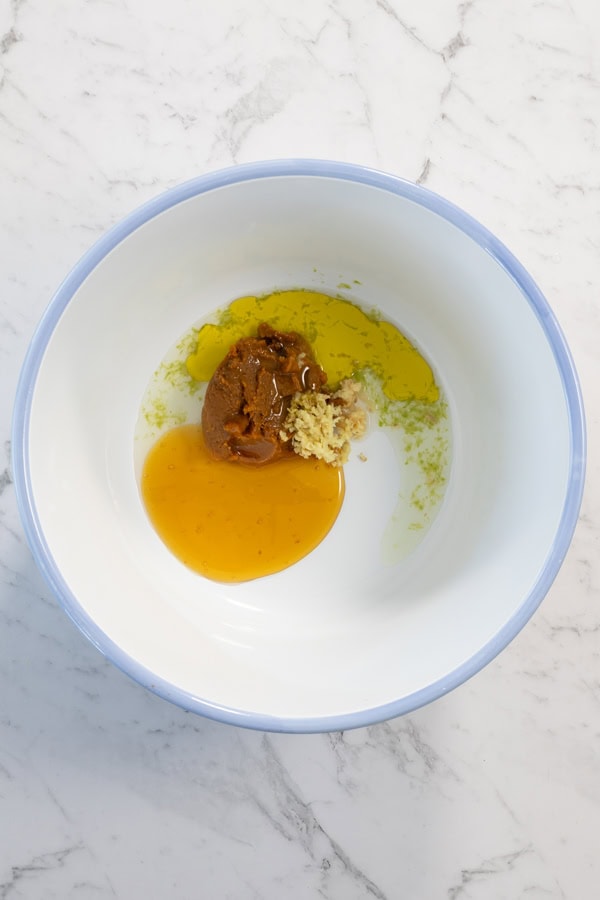 Miso paste, grated ginger, honey, and oil in a white bowl, ready to be mixed.