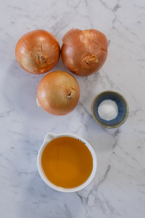 Three brown onions, a bowl of salt, and a measuring cup of oil on a marble surface.