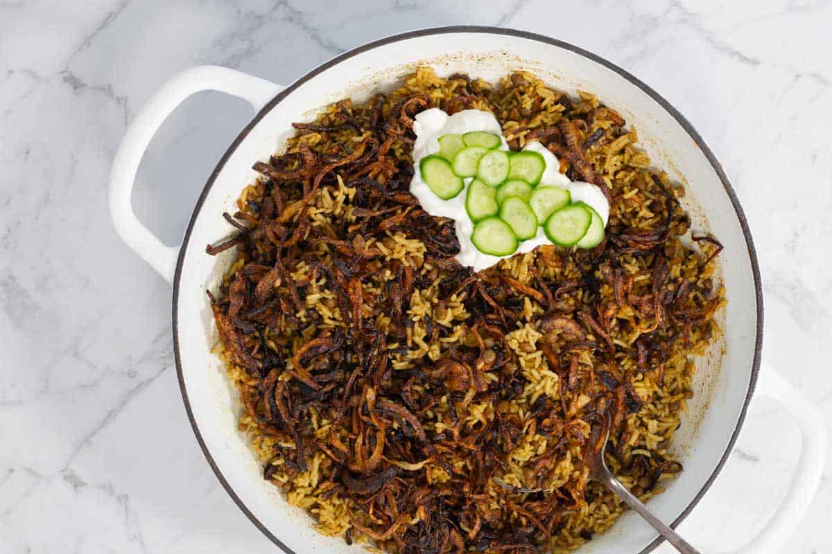 Lentil rice served in a white pot, topped with crispy onions, yogurt, and cucumber slices.
