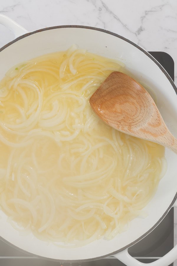 Sliced onions cooking in oil in a white pan with a wooden spoon.