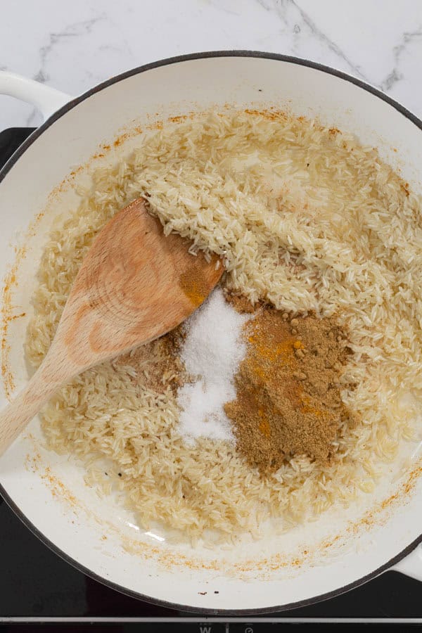Uncooked basmati rice and spices in a white pan with a wooden spoon.