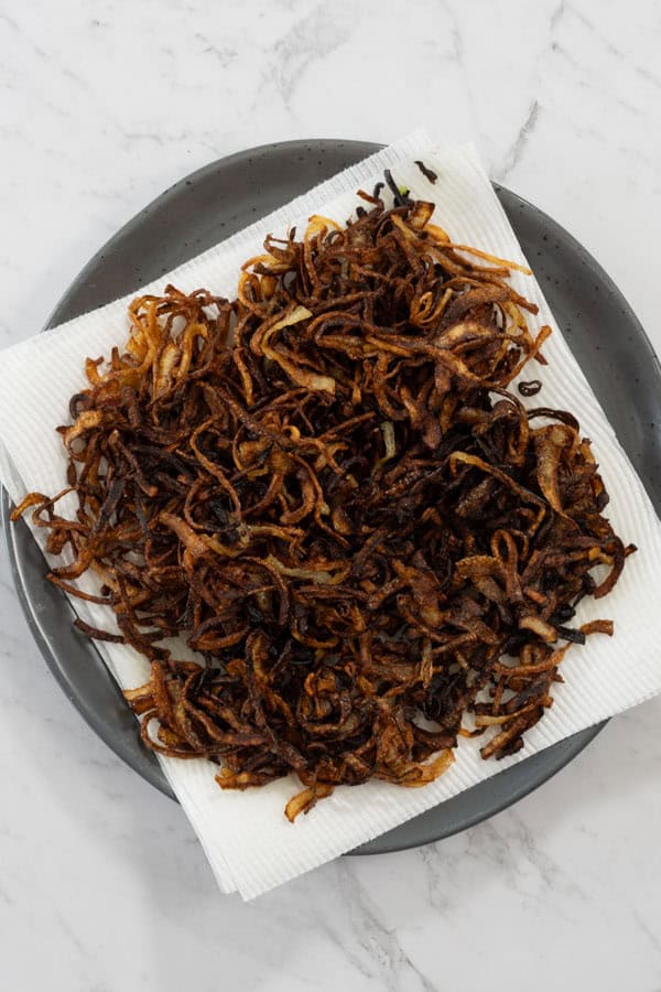 Crispy fried onions draining on a paper towel-lined black plate.