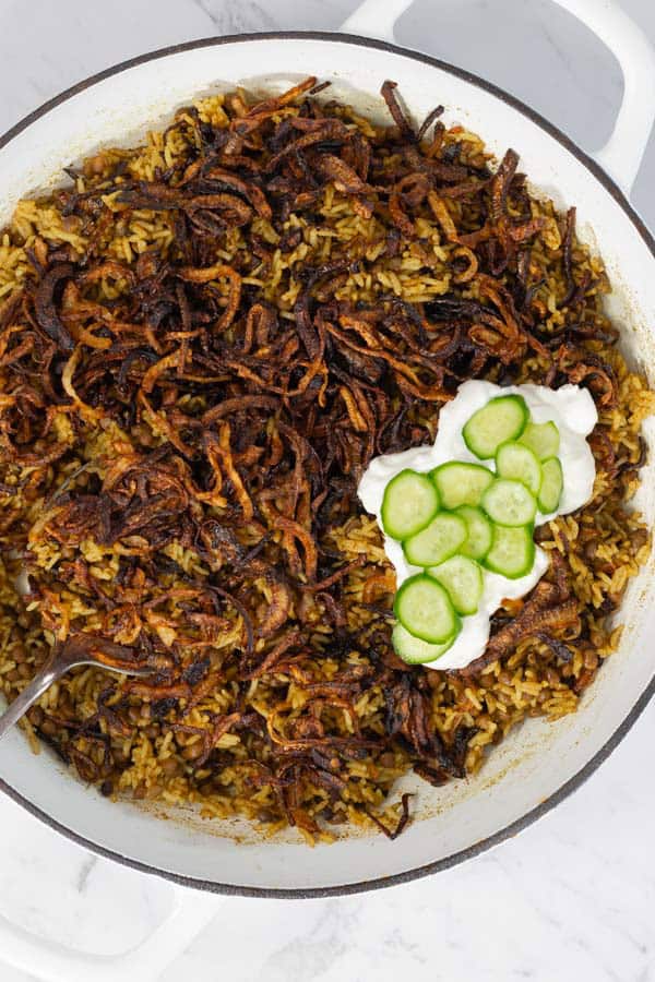 Cooked lentil rice topped with crispy onions, yogurt, and sliced cucumber in a white pot.