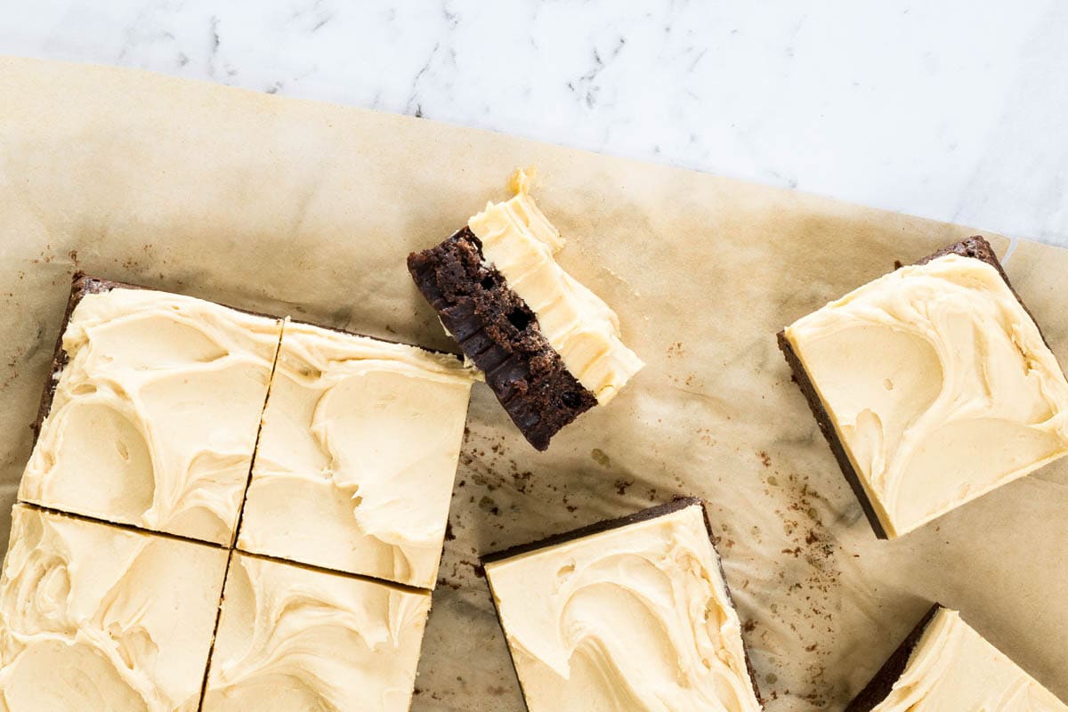 Square-cut dark chocolate brownies topped with a thick swirl of light-tan frosting rest on brown parchment paper for chocolate and peanut butter brownies.