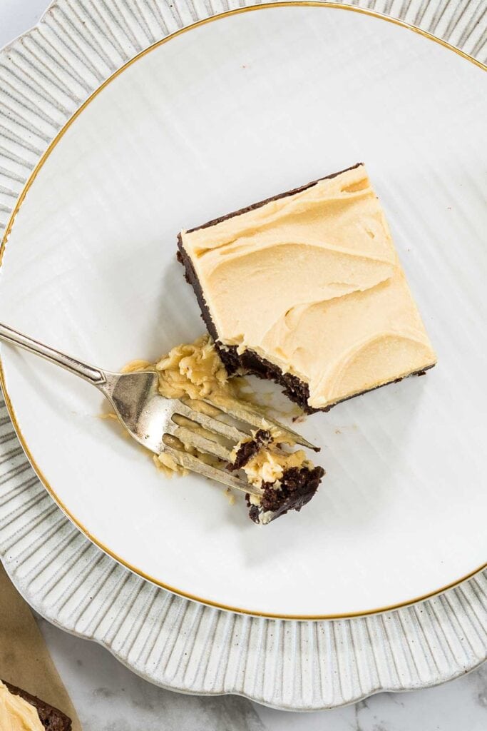 An overhead shot of a single square peanut butter brownie on a white gold-rimmed plate, with a silver fork taking a bite of the dark chocolate base and creamy frosting.