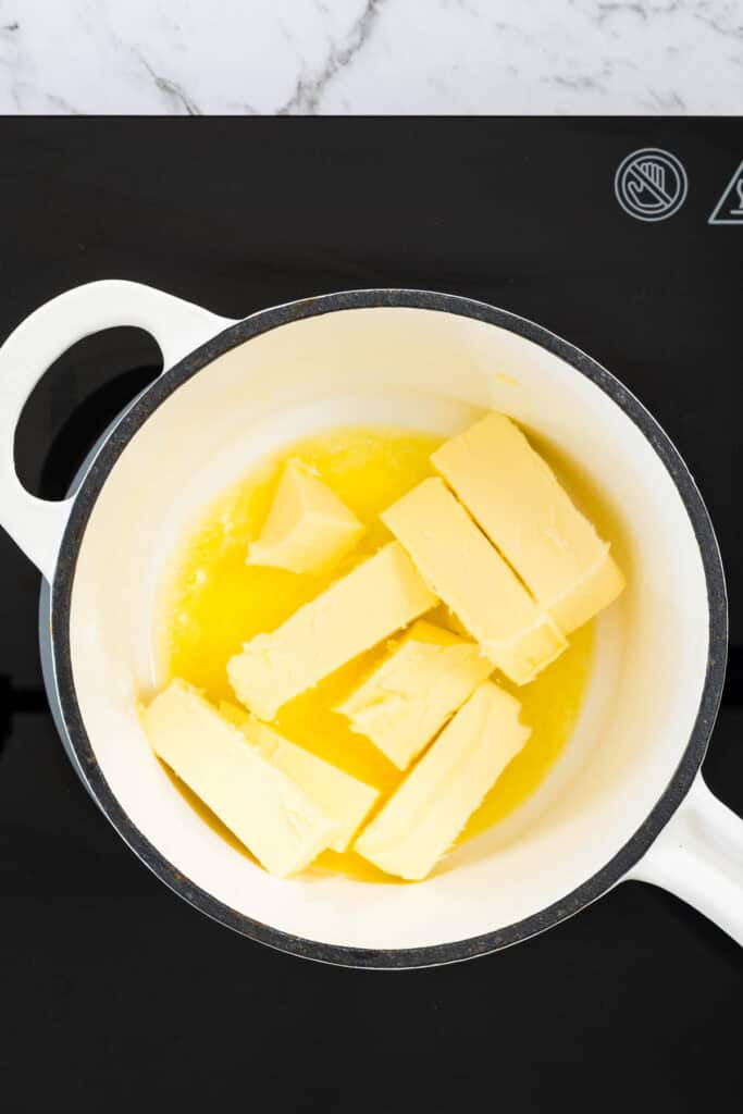 Yellow butter sticks melt into a liquid pool inside a white ceramic pot on a black stovetop for chocolate and peanut butter brownies.