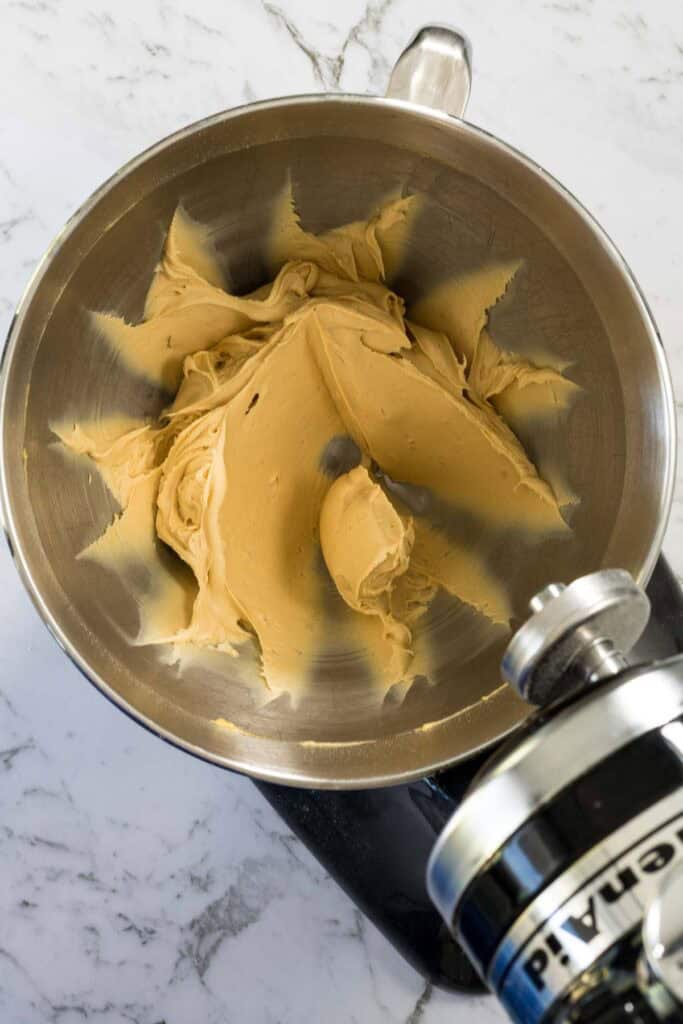 Smooth, pale-brown peanut butter mixture is spread against the sides of a stainless steel stand mixer bowl for chocolate and peanut butter brownies.